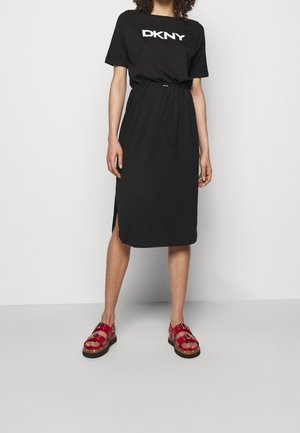 Femme portant une robe midi noire DKNY à manches courtes et taille à cordon, assortie à des sandales rouges à boucle, sur un fond uni.
