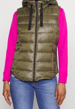 Person trägt eine olivgrüne, gepolsterte ärmellose Weste mit schwarzen Kordeln über einem knallpinken Langarmshirt und blauen Jeans.