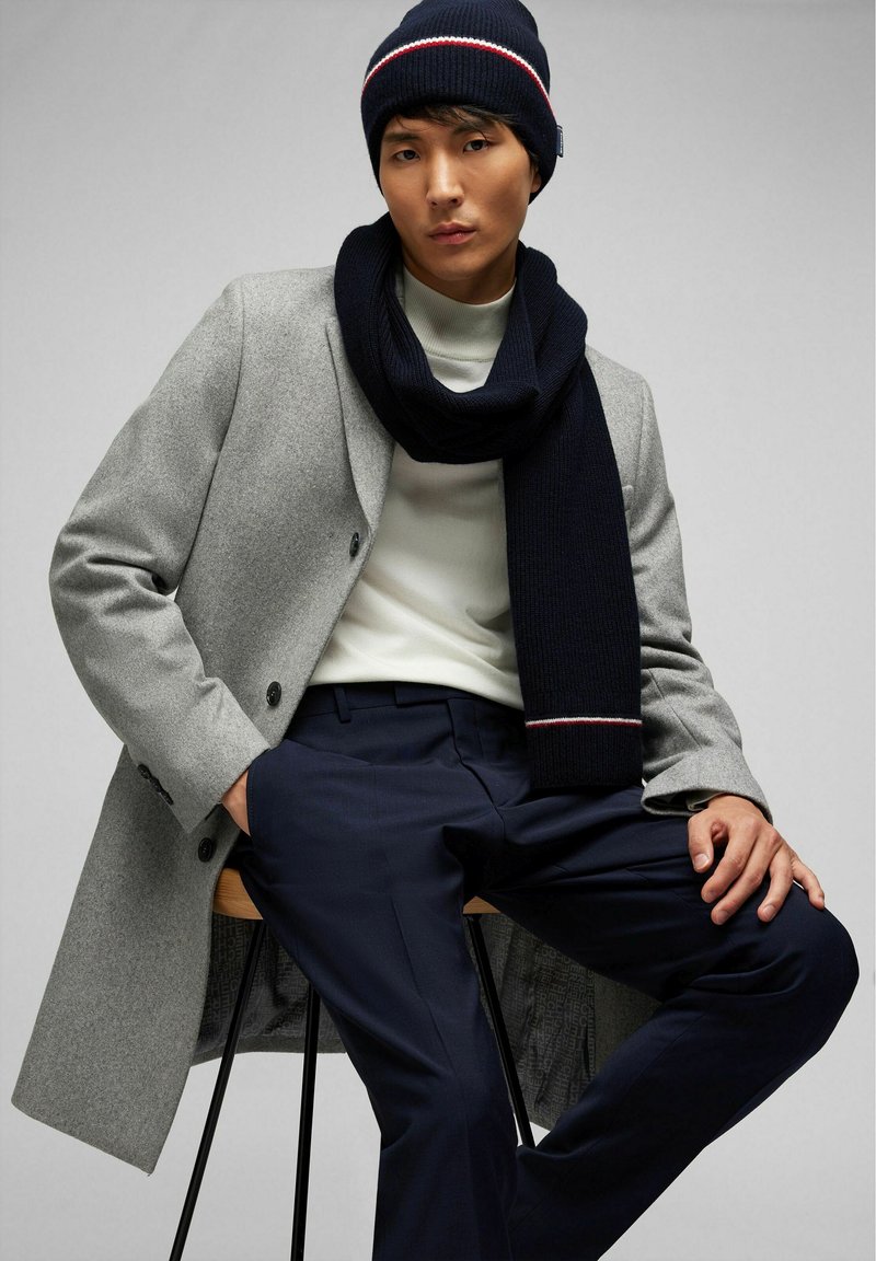 Bonnet tricot marine avec une rayure rouge et blanche, associé à une écharpe marine assortie. Manteau gris clair, pull blanc et pantalons foncés.