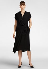 Vestito nero con colletto, maniche corte arrotolate, bottoni frontali e dettaglio in vita annodato. Tessuto morbido con orlo asimmetrico.