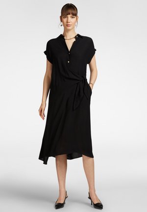 Vestito nero con colletto, maniche corte arrotolate, bottoni frontali e dettaglio in vita annodato. Tessuto morbido con orlo asimmetrico.