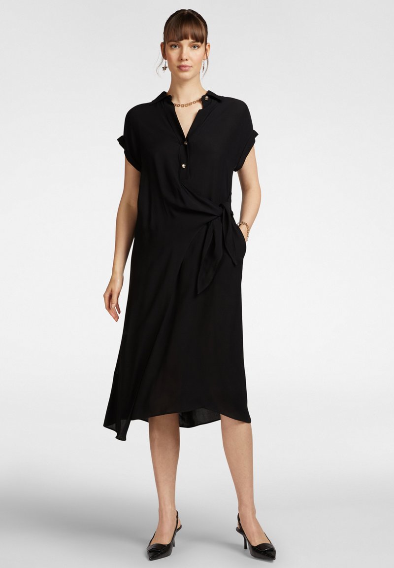 Vestito nero con colletto, maniche corte arrotolate, bottoni frontali e dettaglio in vita annodato. Tessuto morbido con orlo asimmetrico.