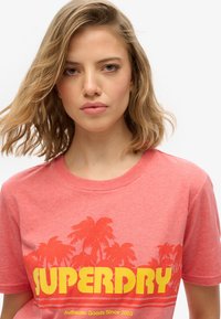 Ung kvinde med skulderlangt blond hår iført en koral Superdry T-shirt med palmetræsgrafik og gul tekst.