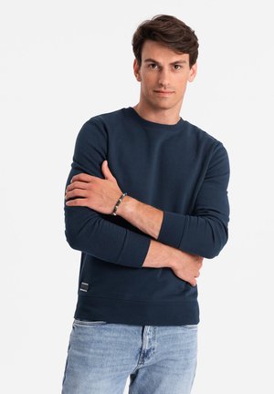 Mörkblå sweatshirt i mjukt tyg med rundad halsringning, ribbade muddarna och en logotypapplikation vid kanten. Kombinerad med ljusblå jeans.