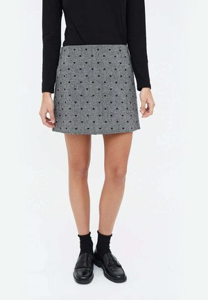 Falda gris con estampado de corazones negros, corte recto y cinturilla ribeteada. El modelo lleva una blusa negra de manga larga y zapatos negros.
