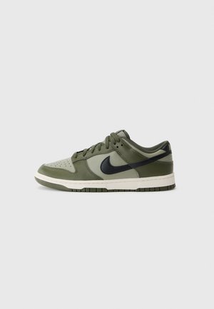 Niski sneaker w kolorze oliwkowo-zielonym i czarnym z sznurowadłami, perforowany czubek i biała podeszwa, widoczny z boku na jasnoszarym tle.