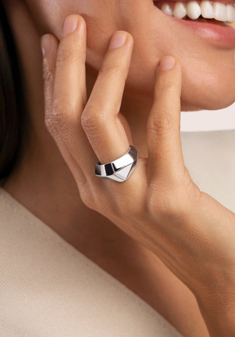 Anello geometrico in argento con superfici lucide e opache, indossato su una mano con texture della pelle naturale. Dettagli di design semplici e moderni.