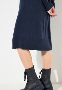 Un vestito in maglione blu navy con orlo a costine, abbinato a stivali alla caviglia in pelle nera con lacci e zip laterale. Tessuto morbido nel complesso.