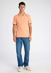 Polo arancione con colletto e polsini a coste neri e bianchi, abbinato a jeans in denim blu e sneakers bianche. Sfondo semplice.