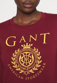 Bordeauxrotes Baumwoll-T-Shirt mit auffälligem gelbem Text "GANT" und einem Wappen mit einer Krone, umgeben von Lorbeerblättern und dem Text "AMERICAN SPORTSWEAR."
