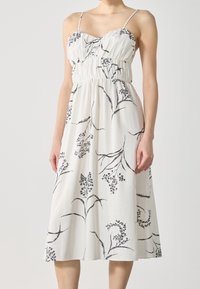 Robe blanche à longueur genou avec des fines bretelles, présentant une taille froncée et un imprimé floral noir, fabriquée en tissu léger.