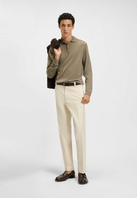 Beige polo met lange mouwen en subtiele textuur, gedragen met crèmekleurige broeken en een bruine riem. Donkerbruine schoenen complementeren de outfit.
