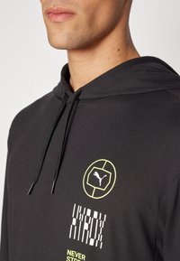 Hettegenser i svart, med en gul sirkulær logo og hvite tekstdetaljer. Materialet ser glatt ut; snorene er svarte.