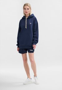 Mörkblå huvtröja med central ficka, dragsko och en liten emblem. Matchas med matchande shorts och vita sneakers. Bomullsmaterial.