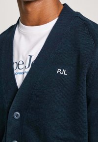 Laivastonsininen neulotun neulepuseron, jossa on resorikuvio, V-aukkoinen malli, edessä napit ja vasemmalla puolella kirjailtu "PJL", puettuna valkoisen T-paidan päälle.