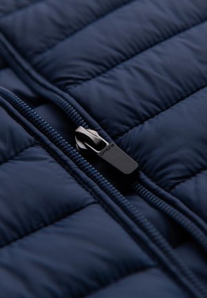 Marineblau gesteppte Jacke mit glatter Textur und einem schwarzen Zipper. Kontrastierender Reißverschluss. Fokus auf den gepolsterten Abschnitten und den Nahtdetails.