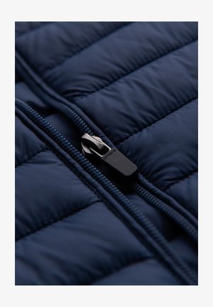 Marineblauwe gewatteerde jas met een gladde textuur, voorzien van een zwarte ritssluiter en een contrasterende rits. Focus op de opgeblazen delen en de stiksels.
