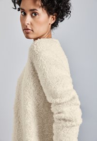 Helle beige, flauschige Strickjacke mit rundem Ausschnitt und langen Ärmeln, aus einem strukturierten, weichen Stoff. Der Hintergrund ist einfarbig grau.