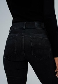 Persona vestida con jeans ajustados negros con bolsillos traseros y una camiseta de manga larga negra, de pie contra un fondo liso.