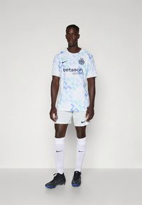 Hvid og lyseblå fodboldtrøje med geometrisk mønster, sort Nike-logo, hvide shorts, sorte fodboldstøvler og hvide sokker med Nike-logo.