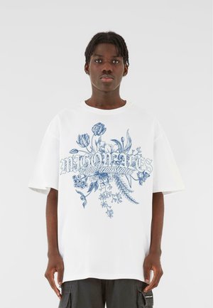 White cotton t-shirt with oversized fit, featuring blue floral design and text "MOONATES" in a bold, decorative font on the front.