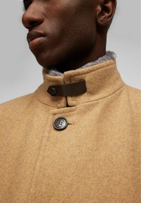 Abrigo de lana color tostado con cuello alto, que presenta una correa de cuero marrón y un cierre de botón. Un suave forro de piel gris asoma visiblemente por el cuello.