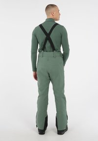Groene buitenbroek met zwarte verstelbare bretels, een getailleerde taille en uitlopende benen met een gestructureerde afwerking en ventilatieopeningen bij de enkels.
