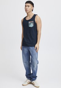 Námornícky modrý tielko s potlačou na vrecku, v kombinácii so svetlomodrými džínsami a bielymi teniskami s hnedými detailmi.