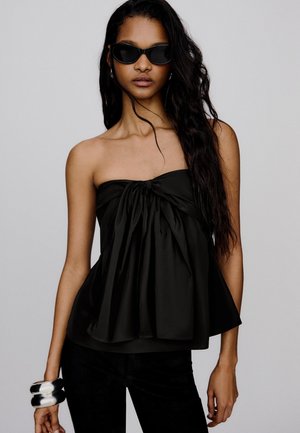 Femme aux longs cheveux noirs portant un haut noir sans bretelles avec un nœud central, un pantalon noir, des lunettes de soleil foncées et de gros bracelets en argent au poignet gauche.