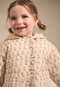 Veste matelassée pour enfants en beige doux avec motif floral, comprenant un devant à volants, une capuche et des boutons ronds. Texture de tissu lisse.