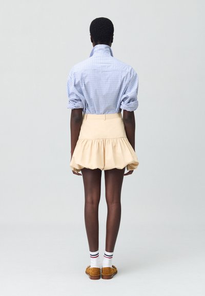 Chemise à carreaux bleus et blancs avec des manches bouffantes, associée à une jupe évasée beige. Chaussettes blanches avec des rayures rouges et bleues, mocassins marron.