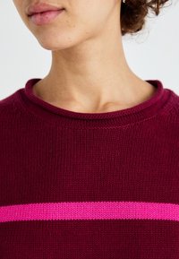 Personne portant un pull en maille bordeaux avec une bande horizontale rose vif sur la poitrine, montrant le cou et le bas du visage.
