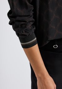 Chemise noire à manches longues avec un motif texturé et des manches froncées. La manche à poignets est dotée d'une bande unie avec deux rayures blanches.