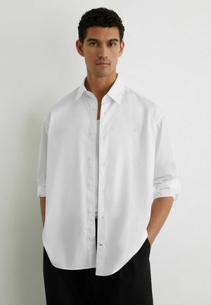 Bărbat cu păr scurt și creț purtând o cămașă albă cu mâneci lungi, desfăcută, peste un tricou alb, stând în fața unui perete simplu.
