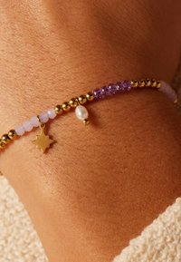 Gouden kralenarmband met lavendel en paarse glasringen, een kleine ster charm, en een witte parelaccent op een gebruinde pols.
