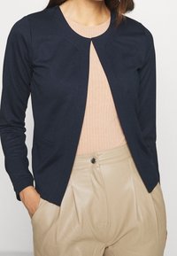 Námořnický cardigan s dlouhými rukávy, kulatým výstřihem a otevřeným předním dílem. Lehká textura, minimální detaily šití. Nesený přes pastelový svetr a béžové kalhoty.