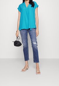 Blusa turchese a maniche corte con scollo a V e nodo frontale, abbinata a jeans chiari strappati e sandali bianchi con cinturini. Inclusa una borsa nera.