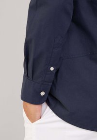 Marineblauwe blouse met lange mouwen en een geworden manchet, met een gladde textuur en een lichte glans. Linkerhandzakdetail zichtbaar, gecombineerd met witte broek.