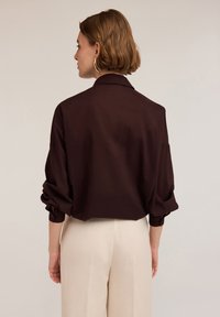 Blusa oversize marrone con colletto e maniche lunghe arrotolate. Tessuto liscio, con taglio dritto e polsini con bottoni.