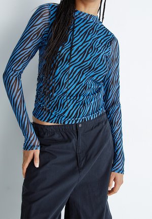 Personne portant un haut à manches longues bleu et noir à rayures zèbre avec des manches transparentes et un pantalon noir ample devant un fond uni.