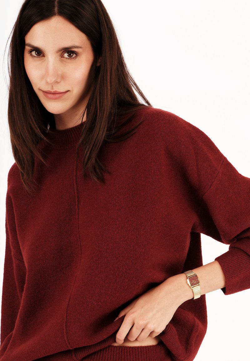 Pull en maille bordeaux avec un large col et des épaules tombantes, présentant une texture subtile. Montre dorée avec un cadran rectangulaire au poignet.