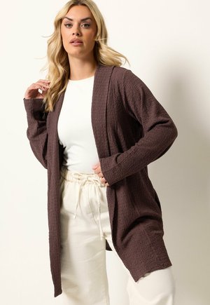 Lyshåret kvinde iført en brun åben cardigan med struktur over en hvid top og cremefarvede bukser med snøre mod en ensartet lys baggrund.