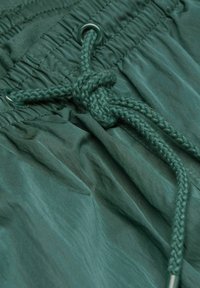 Shorts à cordon de serrage en tissu vert profond, présentant une surface texturée, une taille élastique et un détail de lien tressé avec des œillets en métal.