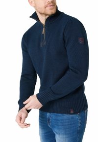 Marineblauer Strickpullover mit einem halbseitigen Reißverschlusskragen, gerippten Bündchen und Saum sowie einem Patchdetail am Ärmel. Hergestellt aus strukturierter Stoff.