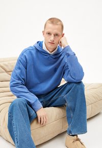 Felpa blu con tasca frontale e logo discreto, abbinata a jeans azzurri chiari. Il modello è seduto su un divano beige con texture.