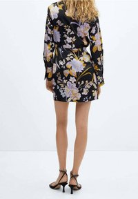 Robe fleurie noire avec des fleurs violettes et jaunes, manches longues et taille cintrée. Complétée par des chaussures à talons hauts noires à brides.