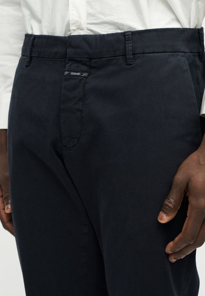 Gros plan sur une personne portant un pantalon bleu marine foncé et une chemise blanche, montrant la ceinture, les passants et la main droite sur le côté.
