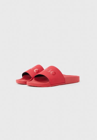 Paire de sandales rouges à enfiler avec larges brides et lettrage "HUGO" en relief sur fond blanc uni.
