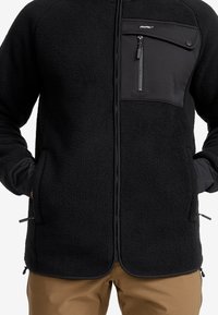 Svart fleecetröja med dragkedjeförsedd bröstficka, elastiska ärmavslut och sidofickor. Mjuk textur med kontrasterande design på frampanelen.