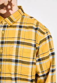 Camisa de cuadros amarilla con grandes patrones en negro y azul, cuello abotonado y un único bolsillo frontal con un discreto detalle de marca.